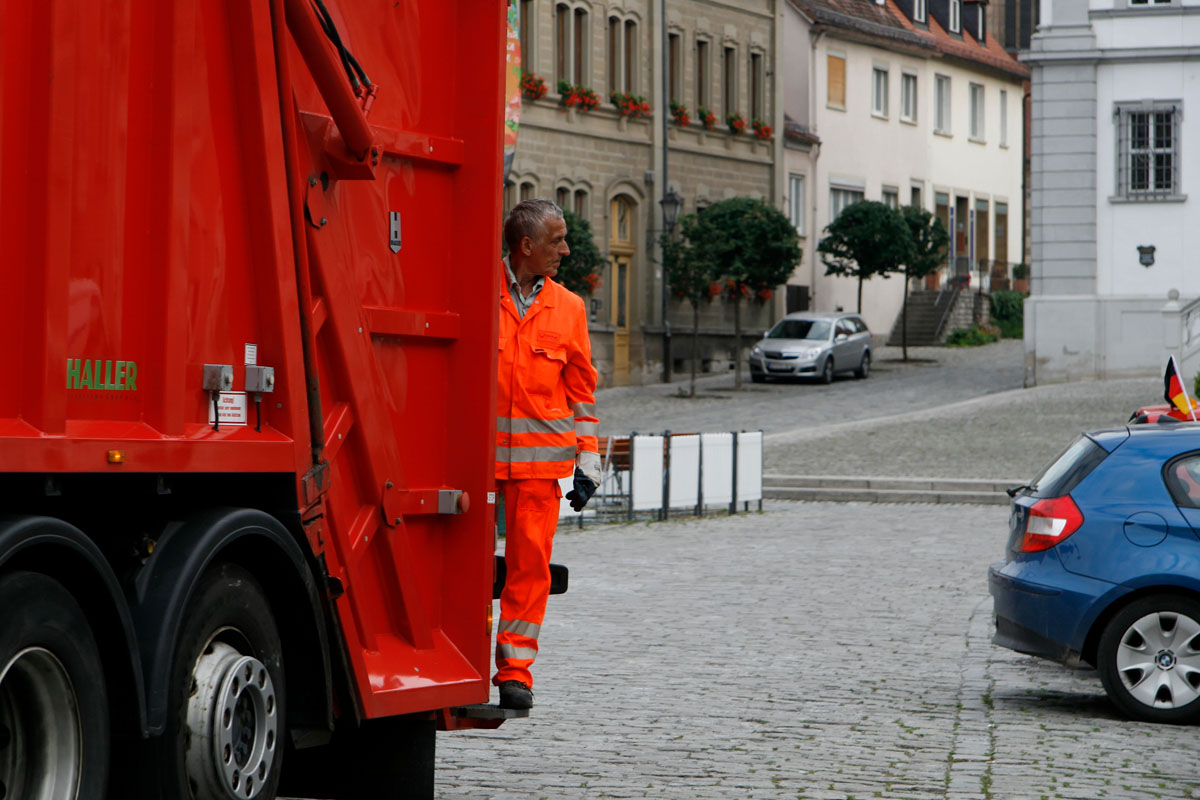 Die Müllabfuhr unterwegs in einer fränkischen Stadt: Müllwerker, Müllfahrzeug, Hecklader, Schüttung, orange Arbeitskleidung, Kopfsteinpflaster