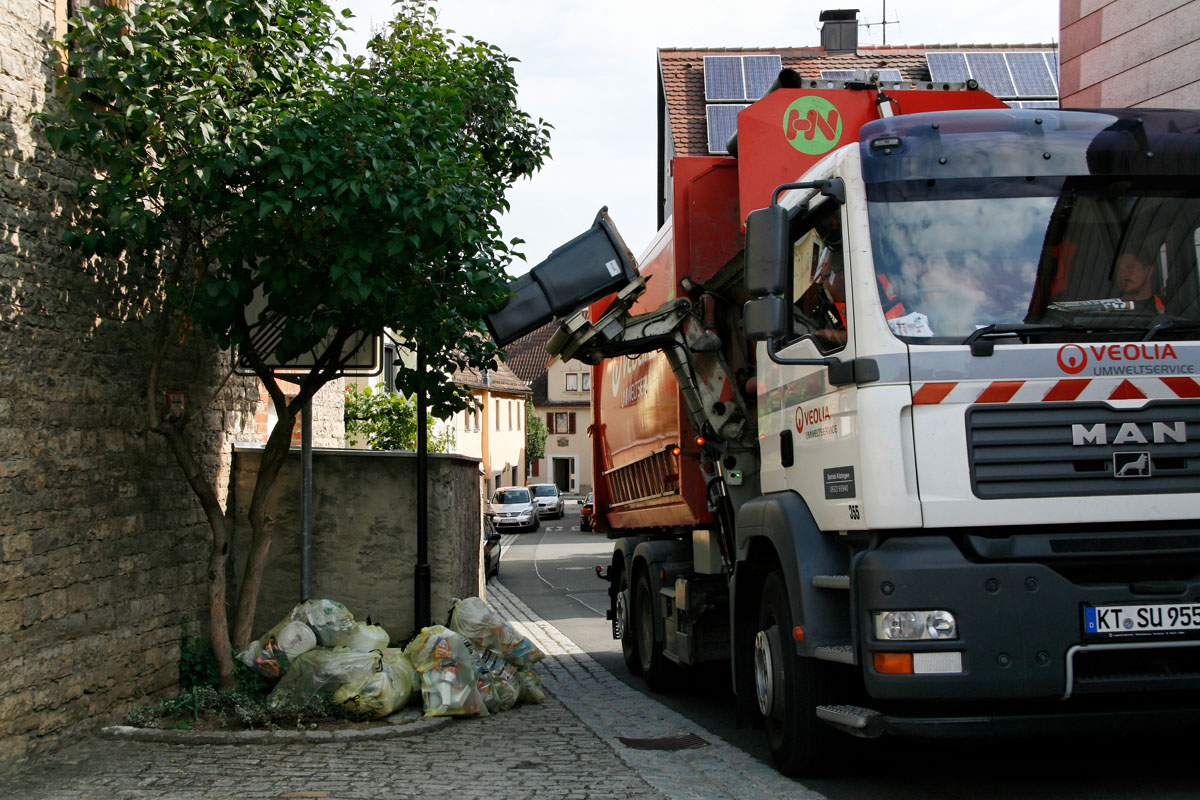 Müllabfuhr mit dem Seitenlader: Leerung einer Biotonne, Gelbe Säcke liegen am Straßenrand zur Abholung bereit
