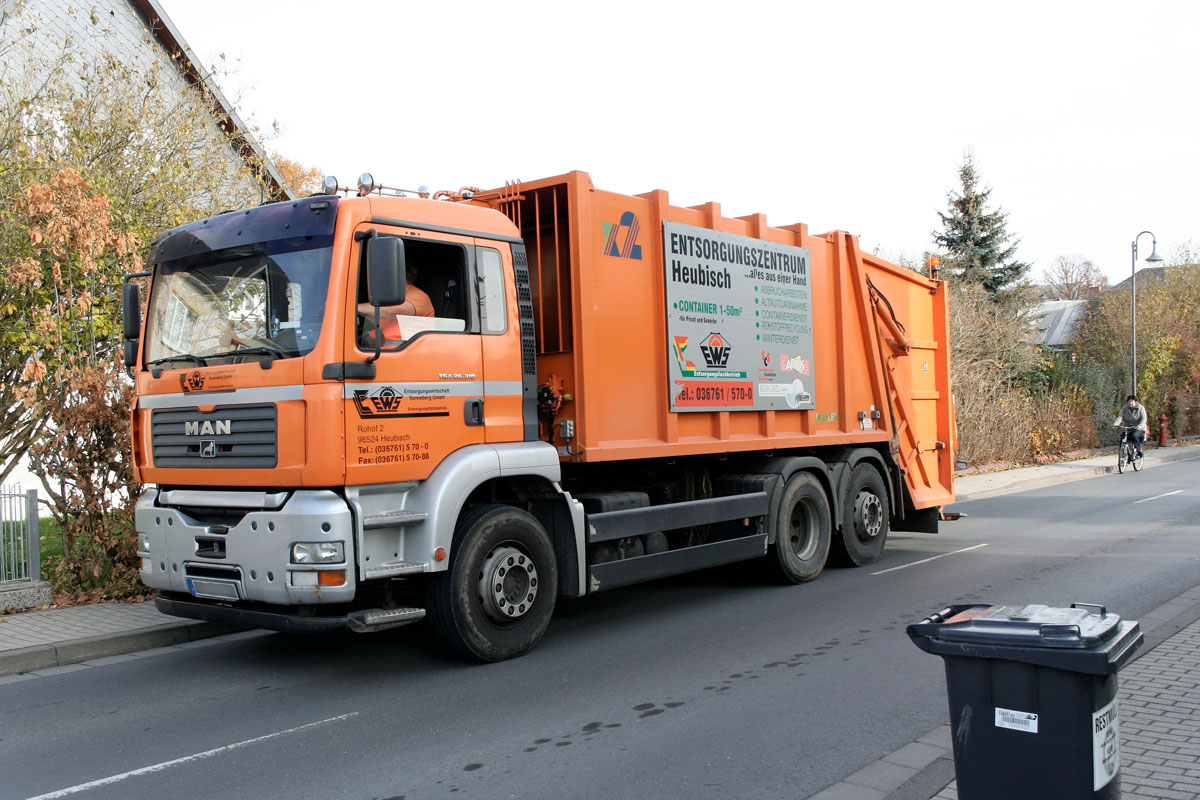 Oranges Hecklader-Müllfahrzeug, graue Restabfalltonne, Straße, Gehweg, Radfahrer, Ortschaft, Haus, Häuser