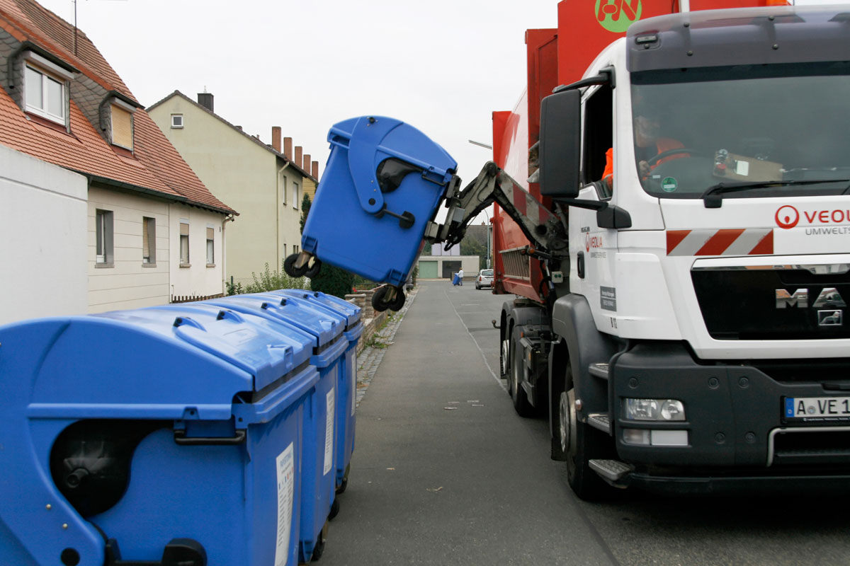 Seitenlader-Müllfahrzeug leert einen blauen Papiercontainer (1.100 Liter MGB) in einem Stadtgebiet mit Mehrfamilienhäusern