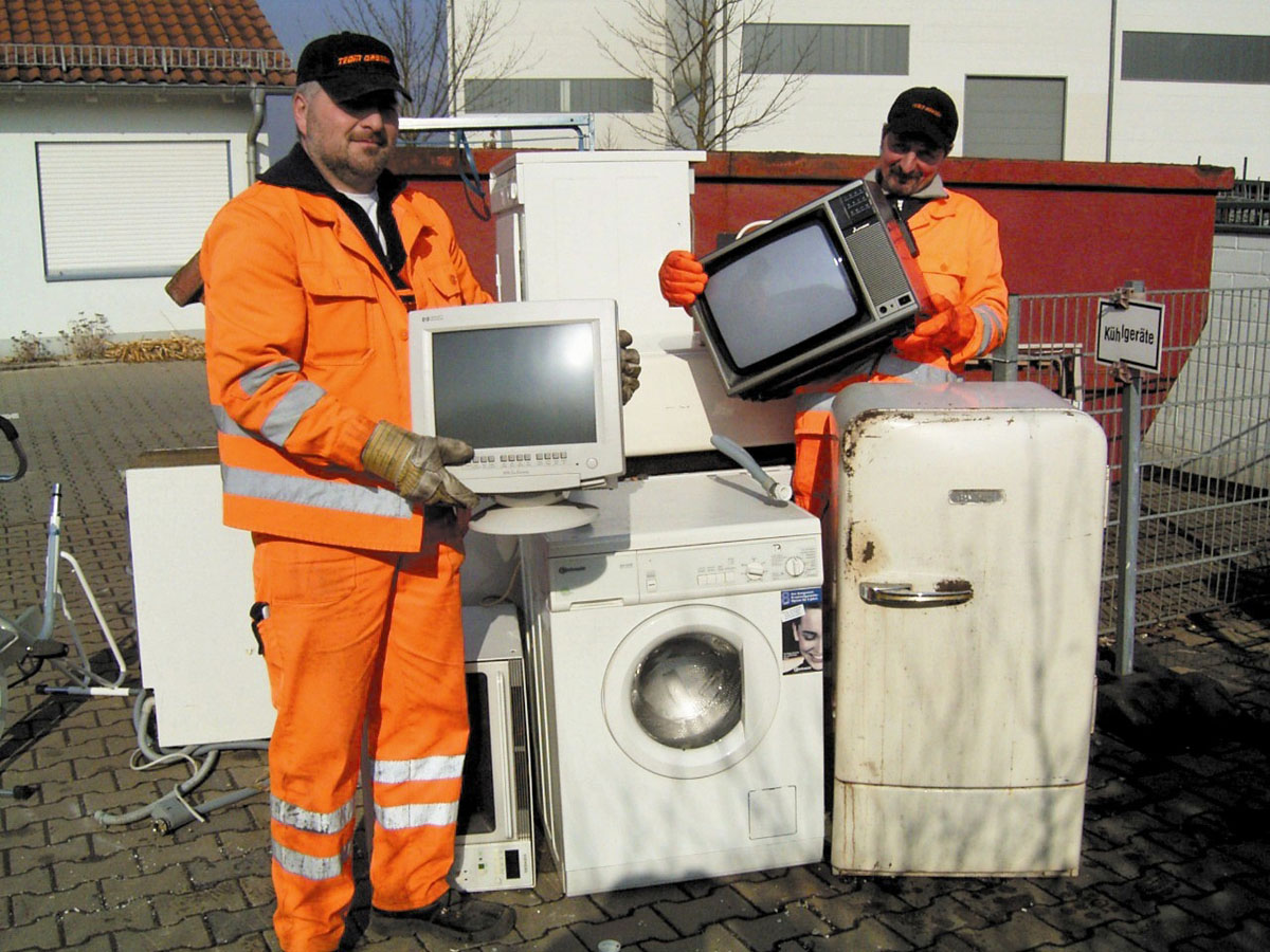 Mitarbeiter eines Wertstoffhofs zeigen gesammelten Elektronikschrott: Fernseher, Computermonitor, Kühlschrank, Waschmaschine, Mikrowelle