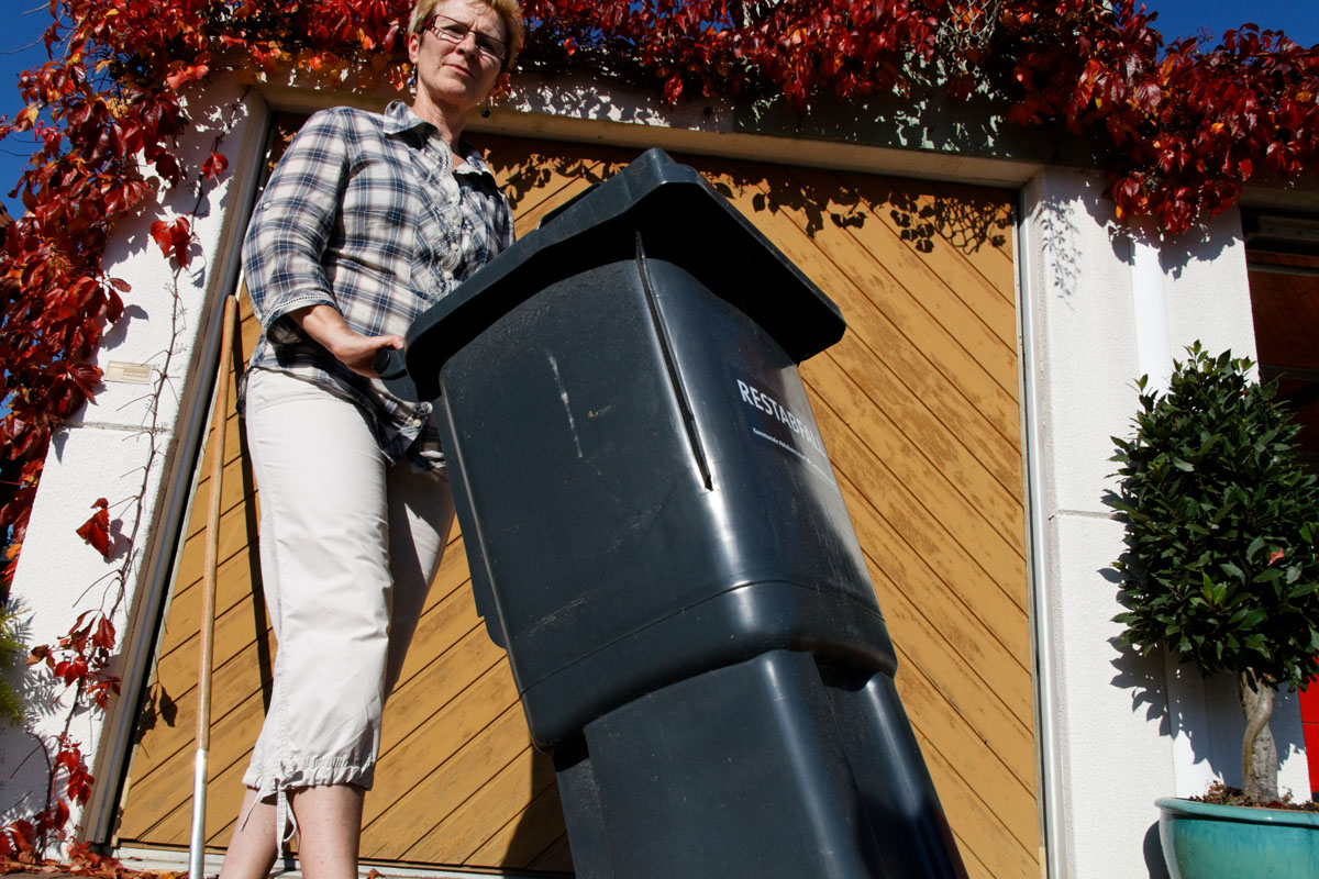 Frau schiebt graue Restabfalltonne zur Müllabfuhr an die Straße