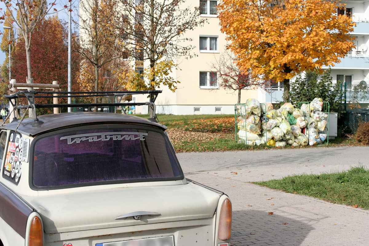 An einer Großwohnanlage (Mehrfamilienhaus, Wohnblock): Gitterrollwagen mit Gelben Säcken, PKW Trabant, Dachgepäckträger