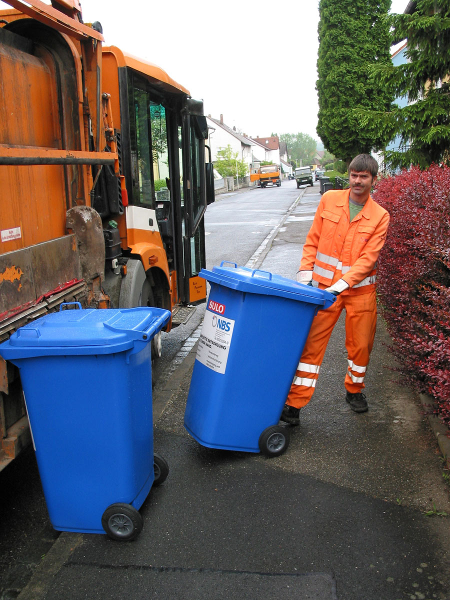 Leerung der Papiertonne mit einem Seitenlader: Müllwerker schiebt blaue Tonnen an das Müllfahrzeug