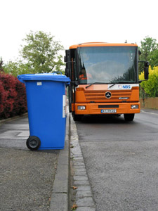 Die Müllabfuhr kommt: Müllfahrzeug nähert sich einer blauen Papiertonne, die am Straßenrand zur Abfuhr bereitgestellt wurde