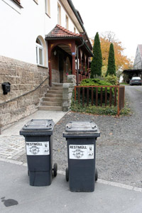 Graue Restabfallbehälter (MGB 120 Liter) stehen am Gehsteig zur Abfuhr bereit: Mehrfamilienhaus, Hauseingang, Fußweg