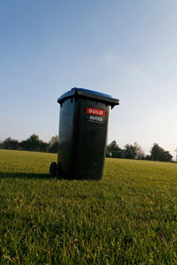 Blaue Papiertonne mit grauem Korpus und blauem Deckel (MGB 240-Liter), Fußballrasen, Sportplatz, Sonne